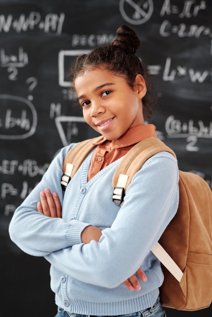 Smiling schoolgirl with backpack poses confidently in front of a classroom chalkboard.